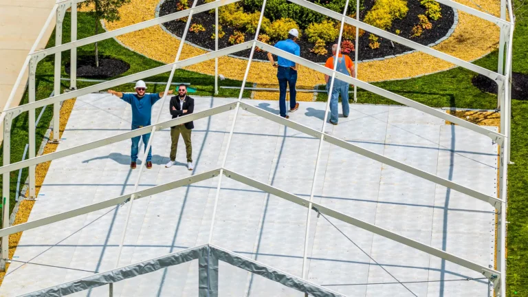 Workers assembling a frame tent with event flooring, preparing for an outdoor event setup.