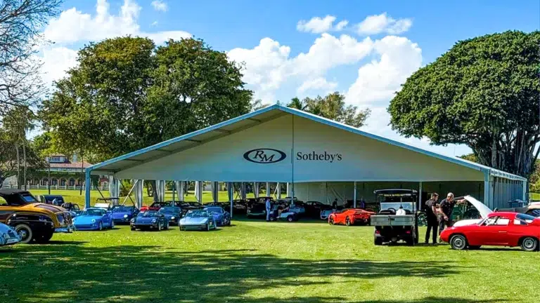 Large frame tent displaying luxury cars at an auction, with event flooring on a grassy field.