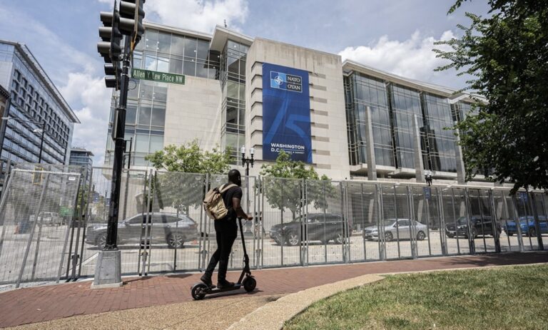 A person riding a scooter passes by crowd control fencing at an event venue with bike rack access, highlighting security and public space management.