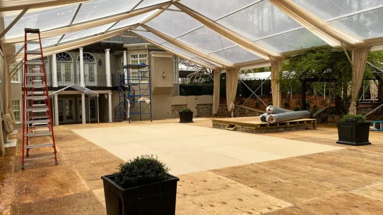 A partially completed event flooring installation inside a clear tent, with plywood sheets laid down and construction work ongoing for a formal event.