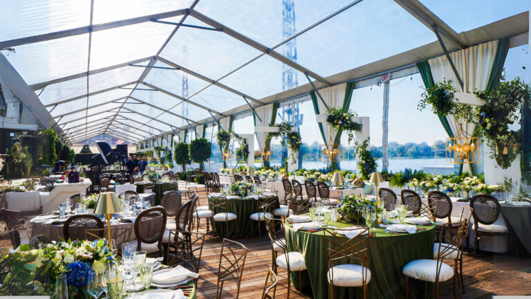 Indoor-outdoor event setup in a clear-frame tent with tables, chairs, floral arrangements, and event flooring overlooking a scenic view.