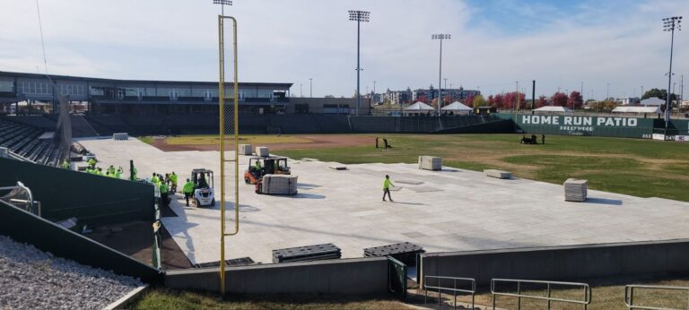 Outdoor stadium field with white turf protection panels stacked, forklifts and event crew installing field floor covers, sports field conversion flooring.