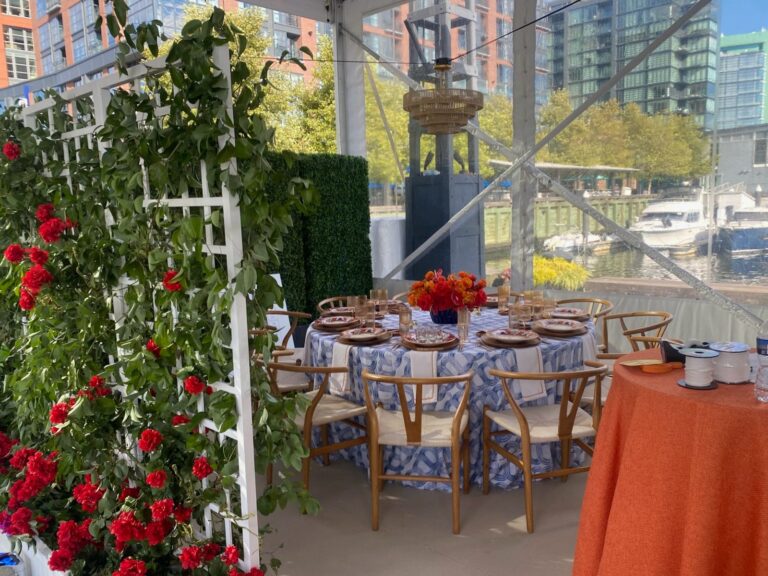 Round table under a clear frame tent, decorated with red roses, wooden chairs, patterned tablecloth, and event flooring.