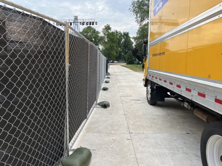 A large fenced event area with delivery trucks and other event setup activities taking place in the background, securing the perimeter of the venue.
