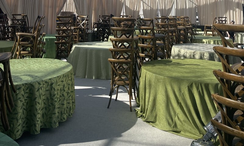 Party tables and chairs for rent set up under a clear tent with round tables, green linens, and wooden chairs