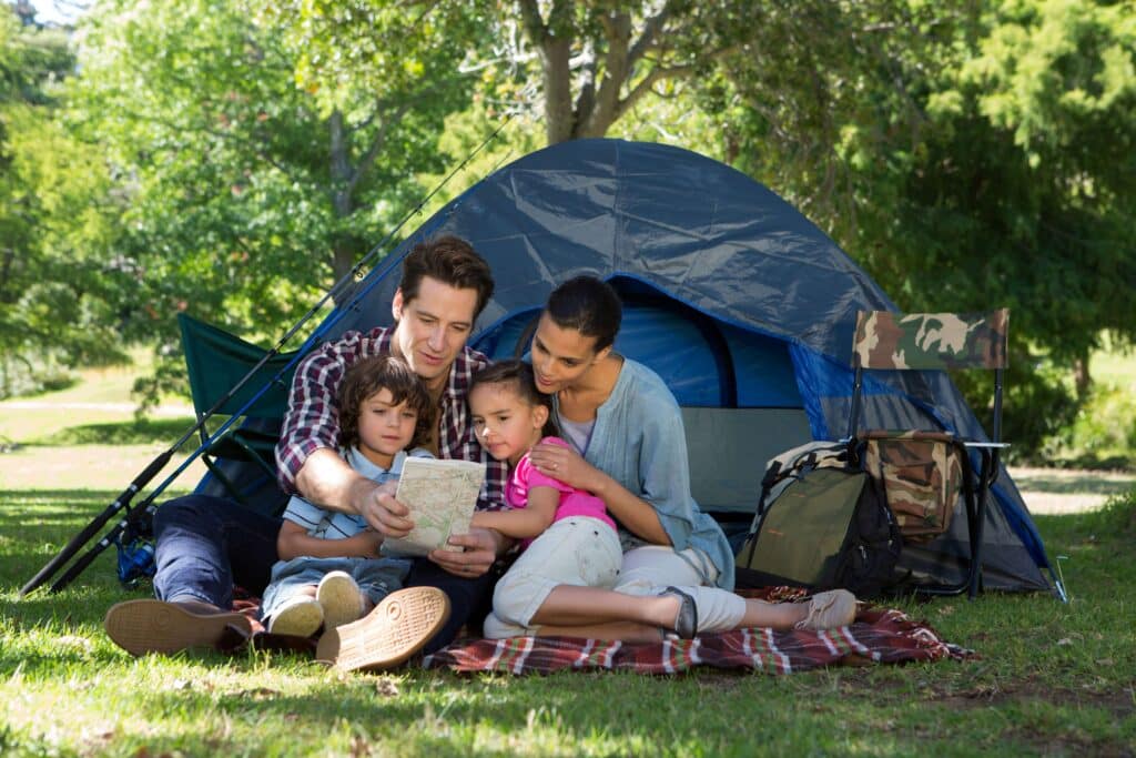 Two adults and two children sitting on a blanket outside their family tent in a park.