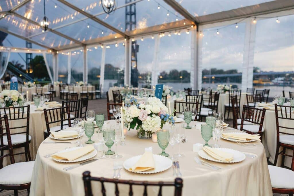 An elegant wedding reception dinner setup with round tables and floral centerpieces inside professional wedding tents featuring clear tops and bistro lighting.