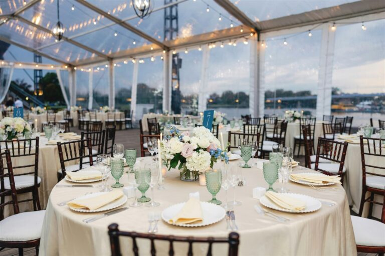 An elegant wedding reception dinner setup with round tables and floral centerpieces inside professional wedding tents featuring clear tops and bistro lighting.