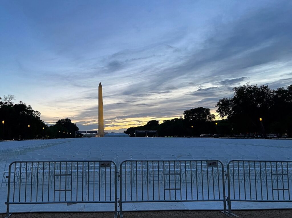 A vast outdoor event space featuring rows of white chairs and professional structure tents set against the sunset near the Washington Monument.
