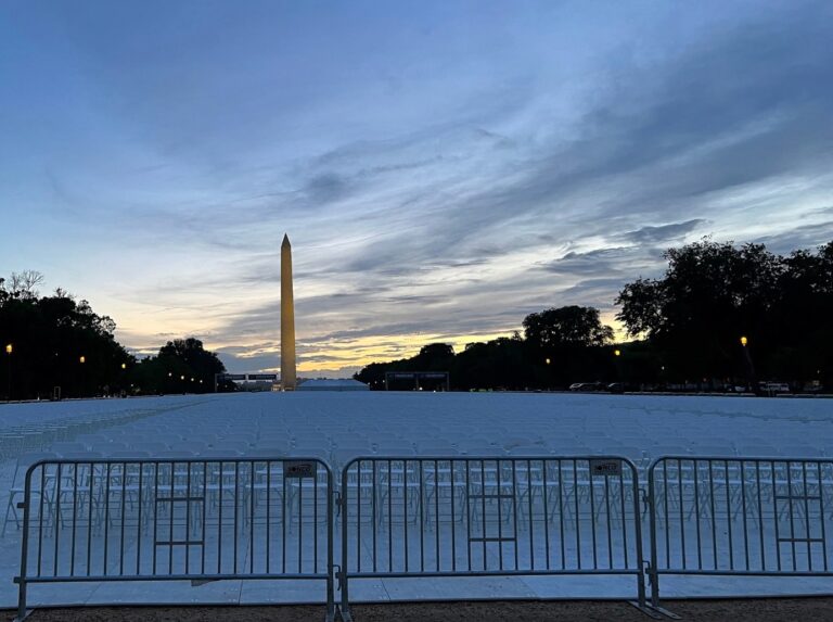 A vast outdoor event space featuring rows of white chairs and professional structure tents set against the sunset near the Washington Monument.