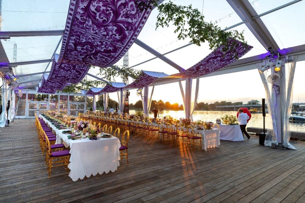 An elegant outdoor dinner setup inside a clear top tent with purple floral fabric accents and sunset views by the river.